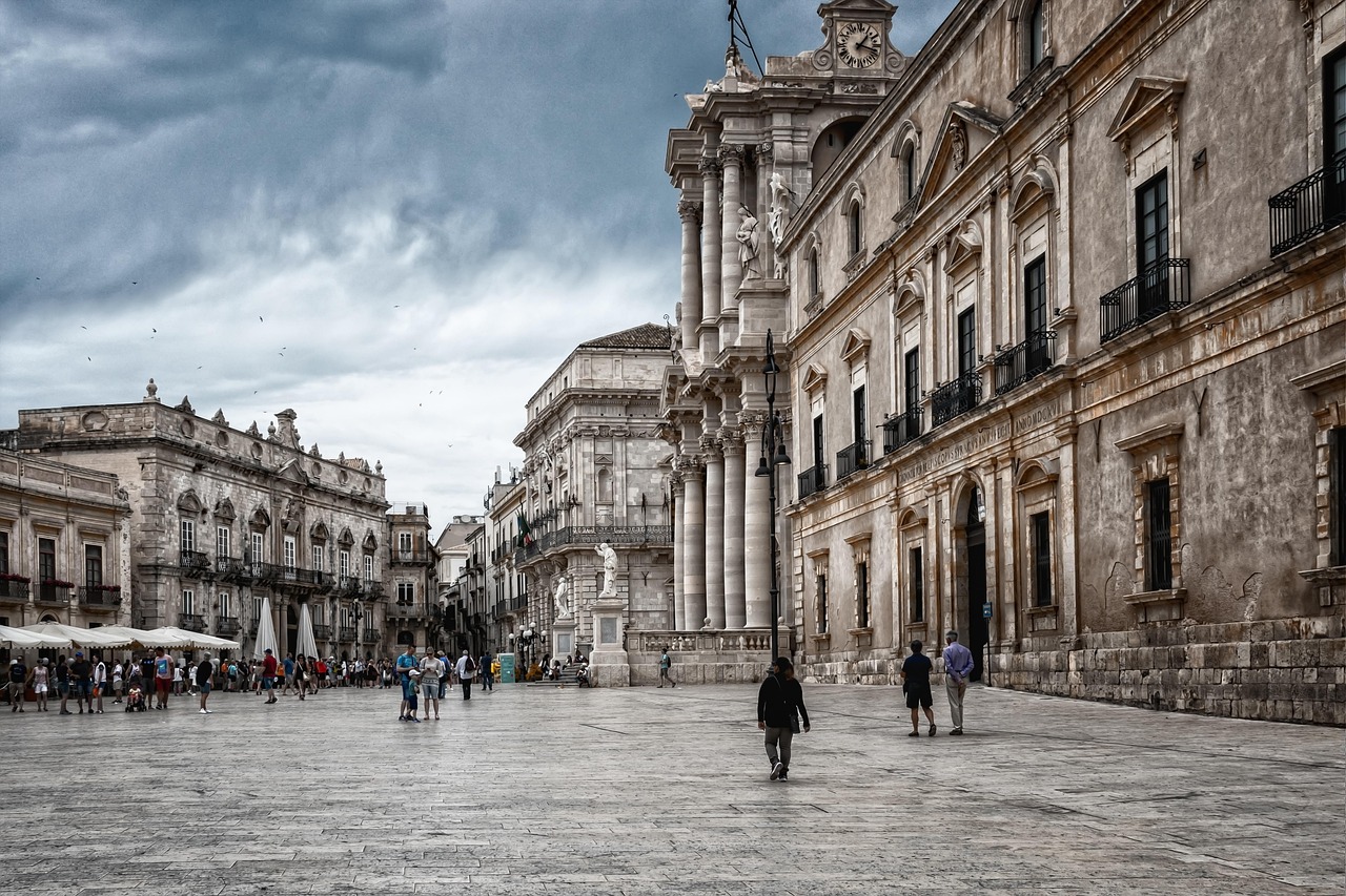 Piazza siciliana affollata, con persone che sorseggiano caffè e chiacchierano. Atmosfera vivace e accogliente.