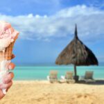 Spiaggia siciliana affollata con gelato artigianale in primo piano, sole e mare cristallino sullo sfondo.